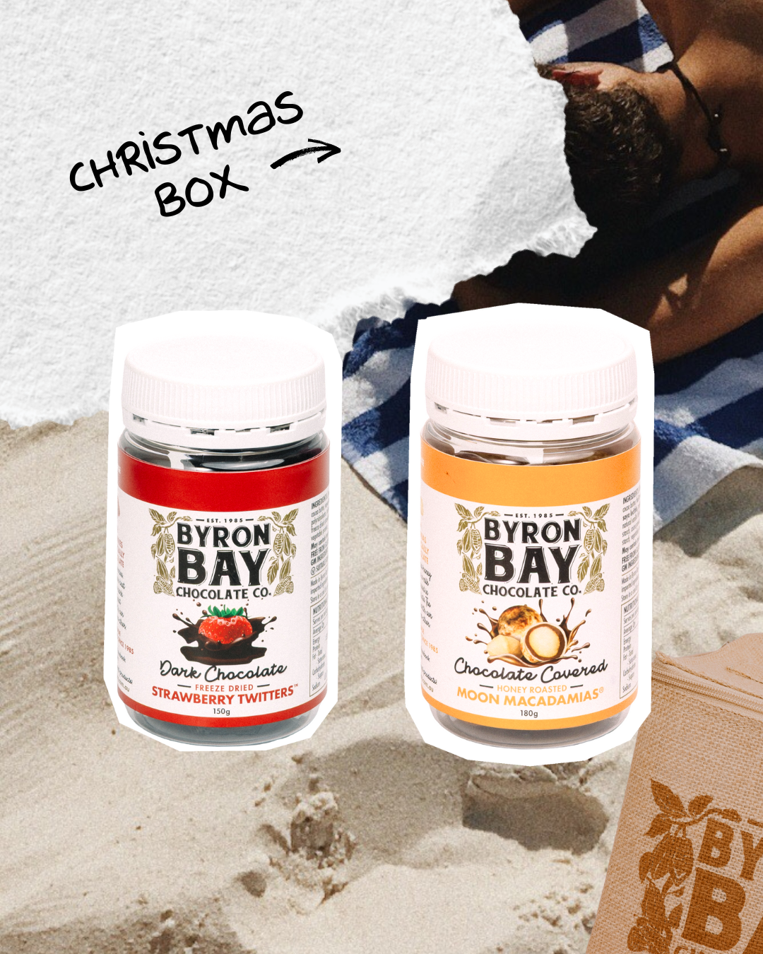 Two Byron Bay Chocolate Co. containers on sand with a Christmas box text overlay.