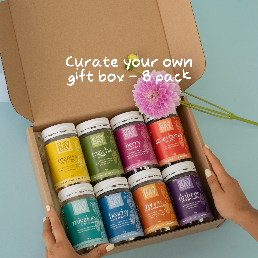 An open cardboard gift box holding eight small Bay Bay essential oil bottles in assorted colors, with a pink flower resting on top. Two hands hold the box edges, and white text across the top reads 'Curate your own gift box - 8 pack'.