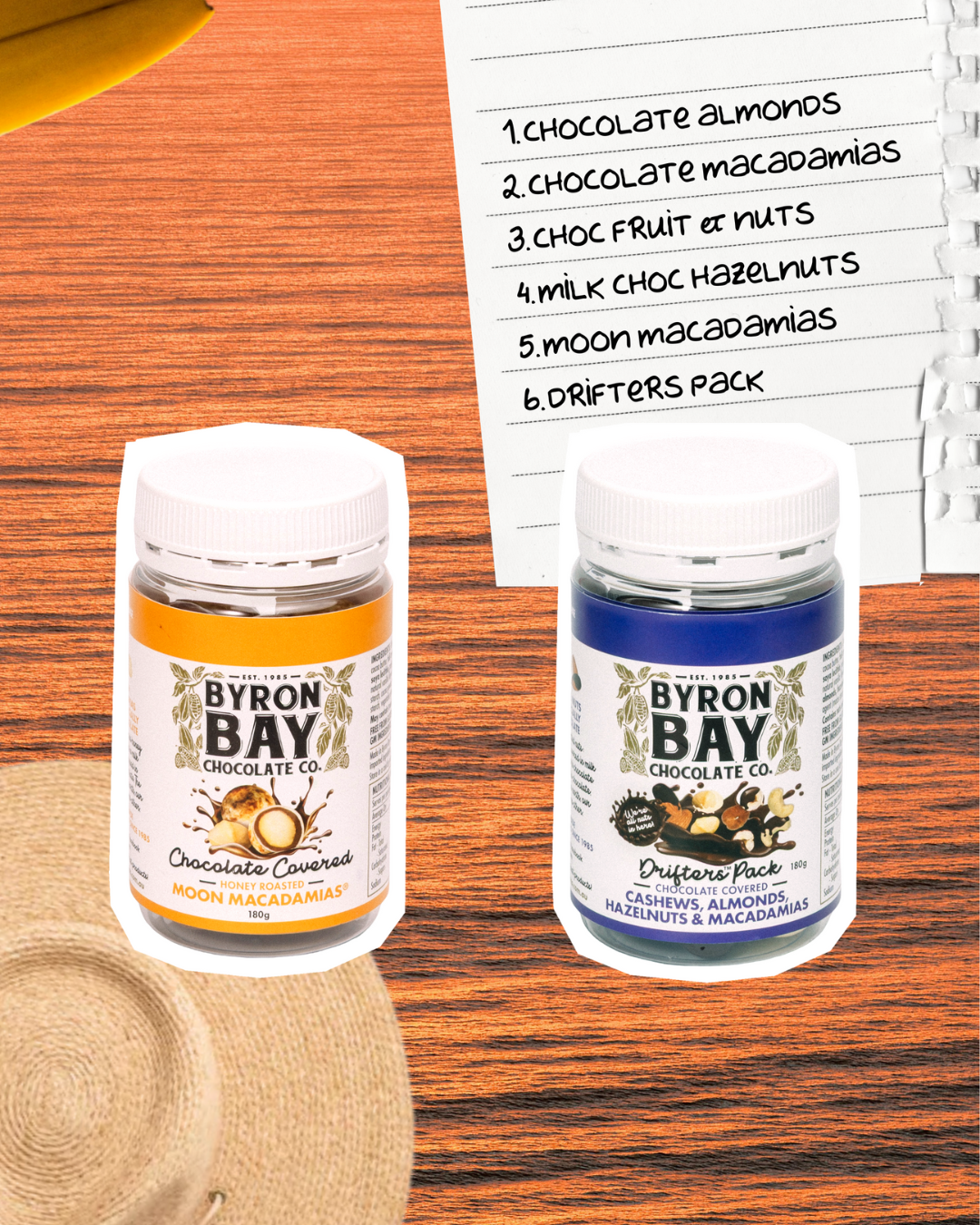 Two jars of Byron Bay chocolate covered nuts on a wooden surface with a list of flavors in the background.